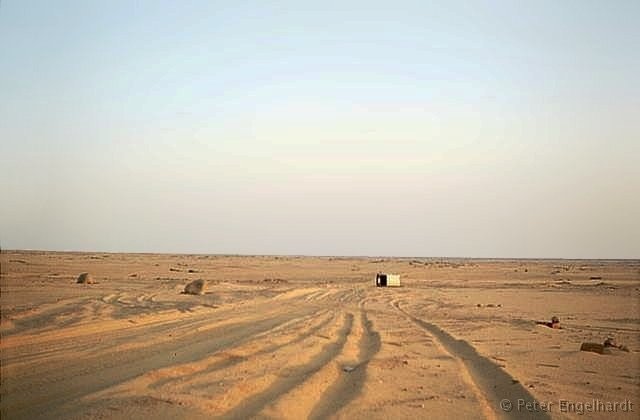 Aufnahme der Wüstenpiste mit einem alten liegengebliebenen Fahrzeug kurz hinter der algerischen Grenze. Rechts erkennt man zwei Wegmarken aus übereinandergelegten Steinen.